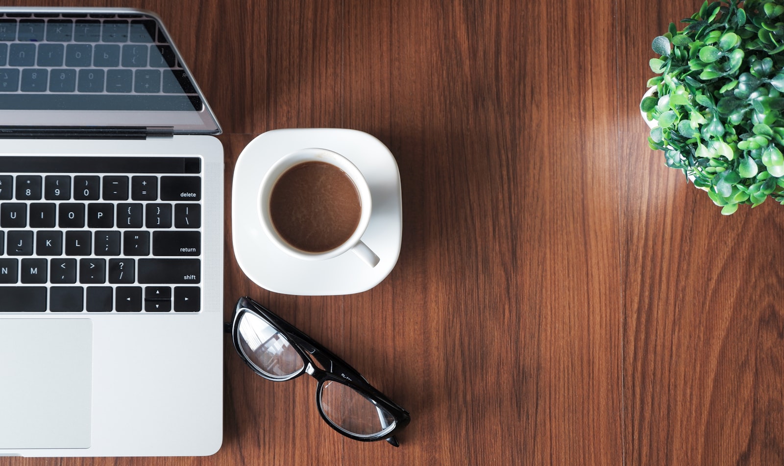 Open laptop, coffee, glasses and a small plant arranged neatly on a warm wooden desk